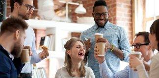 Happy diverse colleagues have fun at lunch break in office, smiling multiracial employees laugh and talk eating pizza and drinking coffee, excited workers celebrate shared win ordering takeaway food.